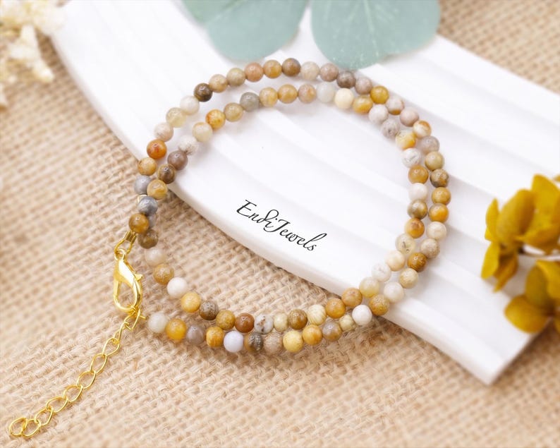 May include: A delicate necklace featuring small, round beads in shades of brown, tan, and cream. The necklace has a gold-colored clasp and chain. The text "End & Jewels" is visible on the white surface.