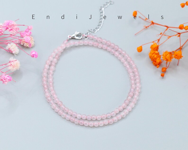 May include: A delicate necklace featuring small, round, pale pink beads arranged in a double-strand design. The piece includes a silver-toned metal lobster clasp and an adjustable chain extension. The text Endi Jewels is visible above the necklace.