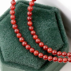 May include: A close-up of a necklace with small, round, reddish-orange beads. The necklace is displayed on a dark green velvet surface, with the beads arranged in two parallel strands. The beads have a glossy finish.