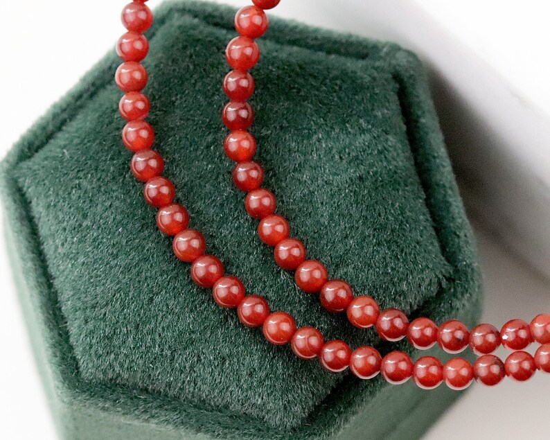 May include: A close-up of a necklace with small, round, reddish-orange beads. The necklace is displayed on a dark green velvet surface, with the beads arranged in two parallel strands. The beads have a glossy finish.