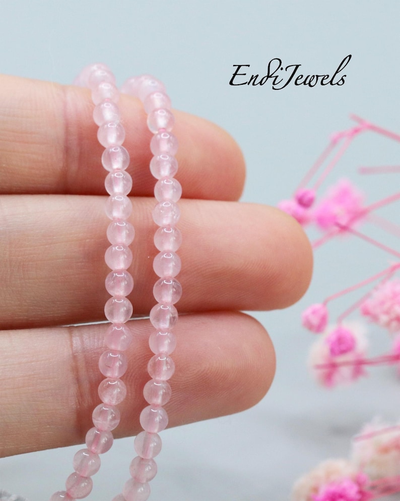 May include: A strand of smooth, round, pale pink rose quartz beads. The text EndiJewels is visible in the upper right corner. The beads are translucent with a polished finish, arranged in a single vertical line against a neutral background.