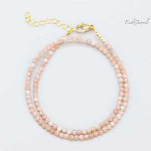 K&ouml;nnte beinhalten: Ein zartes Armband mit doppelter Schnur, bestehend aus hellrosa Sunstone-Perlen und einem goldenen Verschluss. Das Armband ist aus kleinen, facettierten Perlen gefertigt, die einen dezenten Schimmer erzeugen.