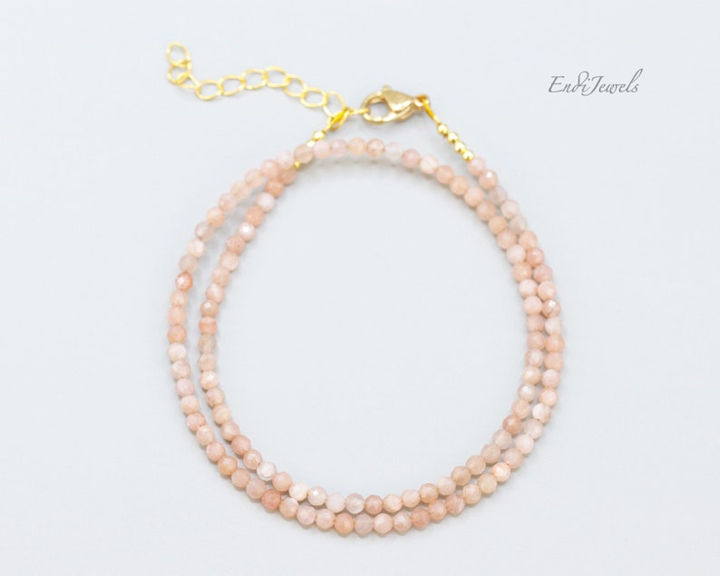 K&ouml;nnte beinhalten: Ein zartes Armband mit doppelter Schnur, bestehend aus hellrosa Sunstone-Perlen und einem goldenen Verschluss. Das Armband ist aus kleinen, facettierten Perlen gefertigt, die einen dezenten Schimmer erzeugen.