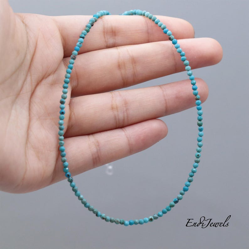 May include: Delicate turquoise bead necklace. The necklace is composed of small, round, light blue beads strung together. The beads are held in a hand against a gray background. The necklace is approximately 16 inches long.