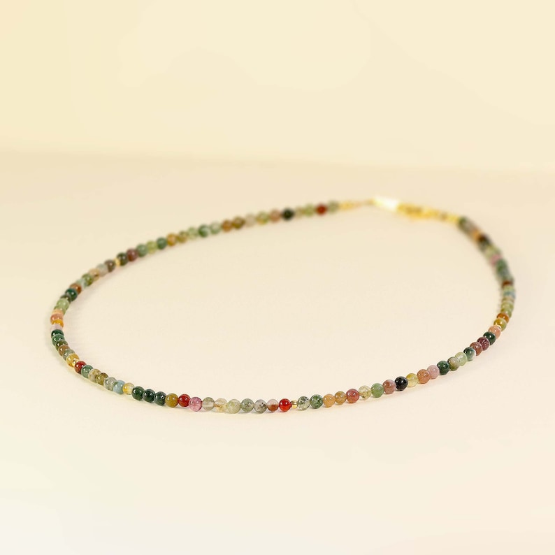 May include: A delicate necklace made of small, multi-colored beads. The beads are a mix of green, brown, red, and pink.