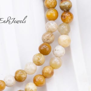 May include: Close-up of two strands of beaded necklaces. The beads are round and vary in color from cream to golden yellow and brown. The necklaces are displayed on a white surface. The text "Endi Jewels" is in the upper left corner.