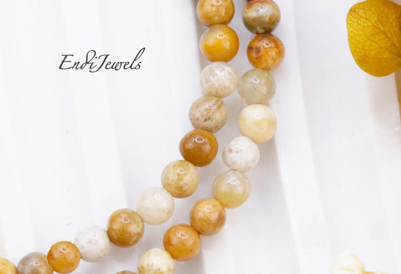 May include: Close-up of two strands of beaded necklaces. The beads are round and vary in color from cream to golden yellow and brown. The necklaces are displayed on a white surface. The text "Endi Jewels" is in the upper left corner.