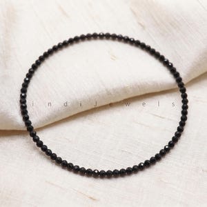K&ouml;nnte beinhalten: Ein kreisf&ouml;rmiger Strang aus kleinen, facettierten schwarzen Perlen, die in einer Ringform angeordnet sind. Der Text "indiJewels" ist in der Mitte des Perlenkreises aufgedruckt. Die Perlen haben eine reflektierende, dunkle Oberfl&auml;che.