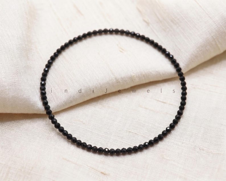 K&ouml;nnte beinhalten: Ein kreisf&ouml;rmiger Strang aus kleinen, facettierten schwarzen Perlen, die in einer Ringform angeordnet sind. Der Text "indiJewels" ist in der Mitte des Perlenkreises aufgedruckt. Die Perlen haben eine reflektierende, dunkle Oberfl&auml;che.