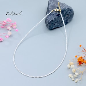 May include: Delicate white pearl necklace with a gold clasp, displayed on a gray surface with a dark gray rock and floral accents. The necklace is finely strung with small, round pearls. The brand name "End&Jewels" is in the upper left corner.
