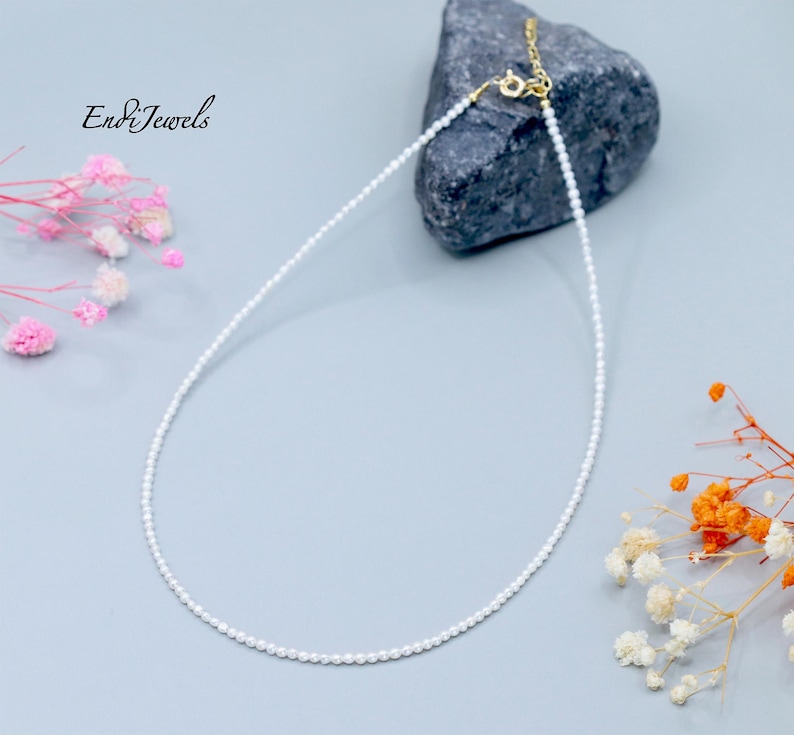 May include: Delicate white pearl necklace with a gold clasp, displayed on a gray surface with a dark gray rock and floral accents. The necklace is finely strung with small, round pearls. The brand name "End&Jewels" is in the upper left corner.