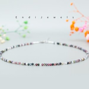 May include: A delicate necklace made of small, multi-colored beads. The beads are a mix of pink, purple, gray, and white. The necklace is strung on a silver chain.
