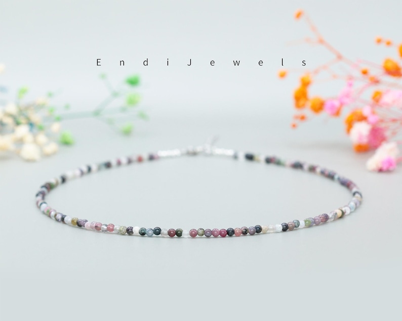 May include: A delicate necklace made of small, multi-colored beads. The beads are a mix of pink, purple, gray, and white. The necklace is strung on a silver chain.