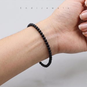 Puede incluir: Una pulsera de cuentas negras se lleva en la mu&ntilde;eca. La pulsera est&aacute; hecha de peque&ntilde;as cuentas negras redondas. La imagen est&aacute; bien iluminada y la pulsera es el punto focal. El nombre de la marca "Endi Jewels" es visible en el fondo.