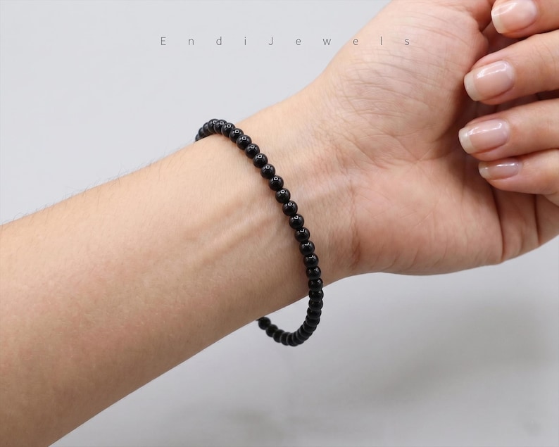 Puede incluir: Una pulsera de cuentas negras se lleva en la mu&ntilde;eca. La pulsera est&aacute; hecha de peque&ntilde;as cuentas negras redondas. La imagen est&aacute; bien iluminada y la pulsera es el punto focal. El nombre de la marca "Endi Jewels" es visible en el fondo.