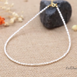 May include: A delicate white pearl necklace with a gold clasp. The necklace is on a brown fabric background.