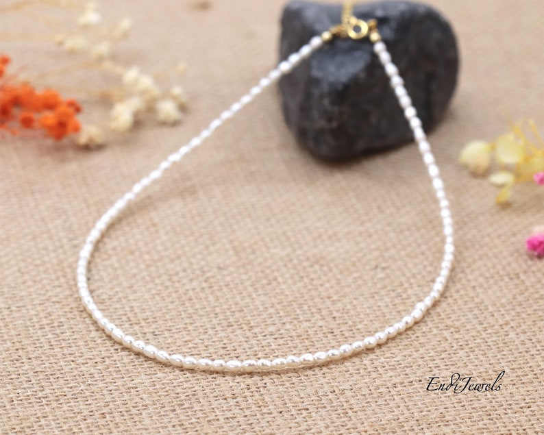 May include: A delicate white pearl necklace with a gold clasp. The necklace is on a brown fabric background.