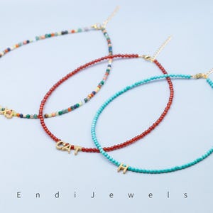 May include: Three beaded necklaces in different colors: multicolored, red, and turquoise. Each necklace has a gold-colored charm with letters or symbols. The necklaces are displayed on a light blue surface. The word "Endi Jewels" is at the bottom.