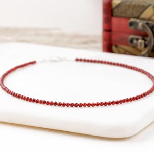 May include: A delicate necklace featuring small, round, red beads strung together. The necklace has a silver clasp and rests on a white surface. The beads are evenly spaced, creating a minimalist and elegant design.