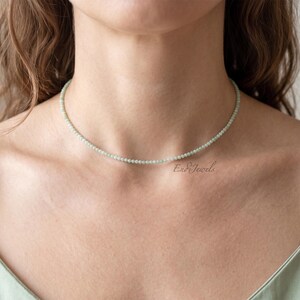 May include: A fine, pale green beaded necklace, a choker, sits on a person's neck. The necklace is made of small, round beads. The person is wearing a light green camisole. The brand name "Ear Jewels" is visible on the lower right.