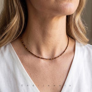 May include: A delicate necklace composed of small, round, brown beads. The necklace is worn around the neck, resting on a white shirt. The text "Endi Jewels" is visible at the bottom of the image.