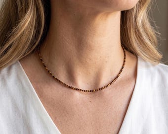 May include: A delicate necklace composed of small, round, brown beads. The necklace is worn around the neck, resting on a white shirt. The text "Endi Jewels" is visible at the bottom of the image.