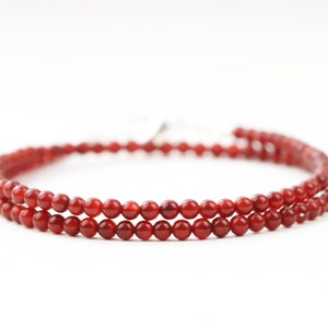 May include: A delicate necklace featuring two strands of small, round, red beads. The beads are strung together, creating a layered effect. The necklace is displayed against a white background, highlighting the vibrant color of the beads.