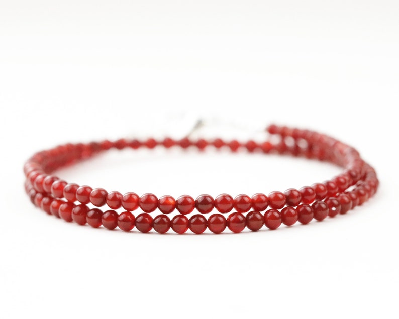 May include: A delicate necklace featuring two strands of small, round, red beads. The beads are strung together, creating a layered effect. The necklace is displayed against a white background, highlighting the vibrant color of the beads.