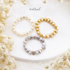 May include: Three pearl rings, one white, one gold, and one silver, on a white surface. The rings are made of small, irregular pearls and have a gold clasp.