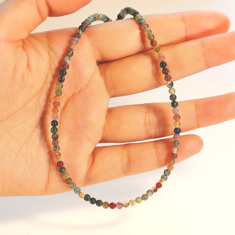 May include: A multi-colored gemstone necklace with small round beads in shades of green, brown, pink, and gold. The necklace is made of natural stones and is strung on a thin gold chain.