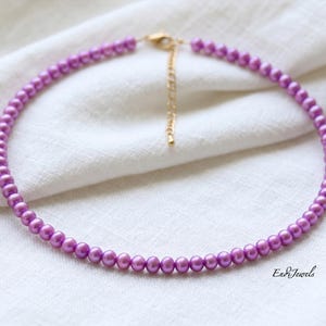 May include: A lavender pearl necklace with a gold-coloured clasp and extender chain. The necklace is made of small, round pearls. It is displayed on a white fabric surface. The brand name "En&Jewels" is visible.