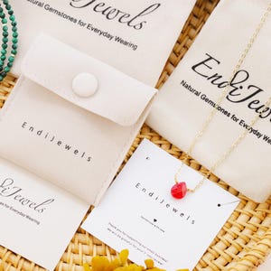 May include: An assortment of jewelry pieces, including a gold chain necklace with a red gemstone pendant and a green beaded necklace, are displayed on a woven tray. The jewelry is packaged in small, off-white pouches and cards with the brand name "Endi Jewels".
