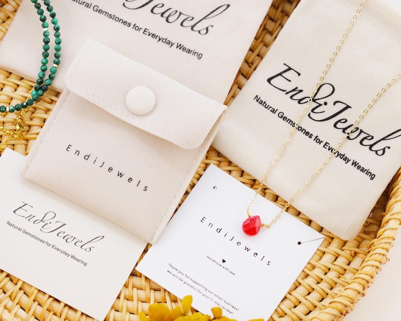 May include: An assortment of jewelry pieces, including a gold chain necklace with a red gemstone pendant and a green beaded necklace, are displayed on a woven tray. The jewelry is packaged in small, off-white pouches and cards with the brand name "Endi Jewels".