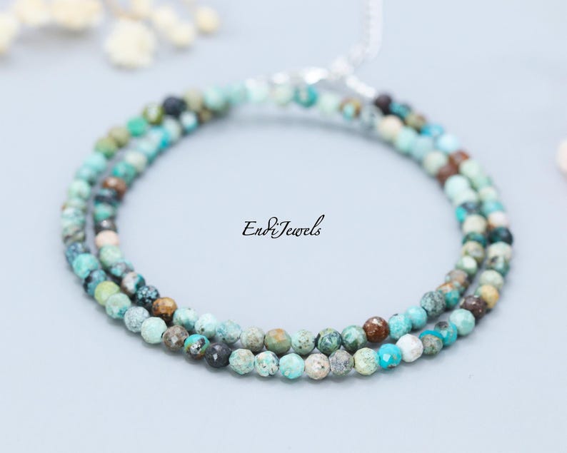 May include: A delicate turquoise beaded necklace with a silver clasp. The necklace is composed of small, round beads in shades of turquoise, brown, and green. The text "EndiJewels" is printed on the right side of the image.