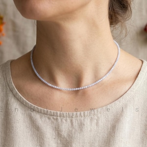 May include: A delicate, light blue beaded necklace. The necklace is composed of small, round beads, creating a subtle, elegant choker style. The jewelry is displayed on a person wearing a neutral-toned top.