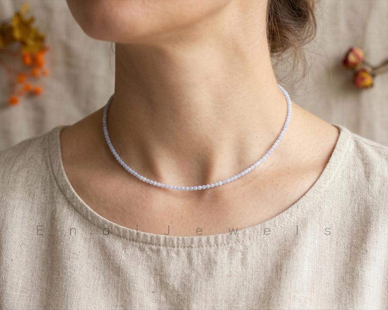 May include: A delicate, light blue beaded necklace. The necklace is composed of small, round beads, creating a subtle, elegant choker style. The jewelry is displayed on a person wearing a neutral-toned top.