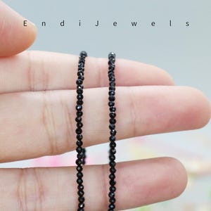 May include: Two strands of black gemstone beads, likely onyx, are held in a hand. The beads are small and round, and the strands are long enough to be worn as a necklace.