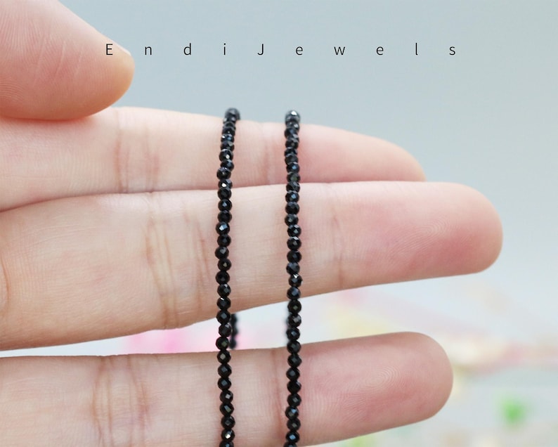 May include: Two strands of black gemstone beads, likely onyx, are held in a hand. The beads are small and round, and the strands are long enough to be worn as a necklace.