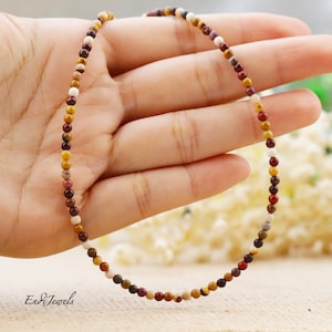 May include: A multi-colored gemstone necklace with small round beads in shades of brown, red, yellow, and white. The necklace is on a hand, and the background is a light brown surface.