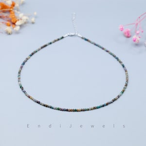 May include: A delicate beaded necklace featuring small, multi-colored beads. The necklace has a silver-toned clasp and chain. The beads are in shades of green, blue, brown, and tan. The necklace is displayed on a light blue surface. The text "Endi Jewels" is at the bottom.