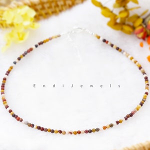 May include: A delicate necklace made with small, multi-colored gemstone beads. The beads are a mix of brown, red, white, and yellow. The necklace is on a white background.