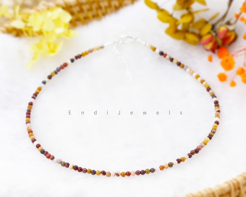 May include: A delicate necklace made with small, multi-colored gemstone beads. The beads are a mix of brown, red, white, and yellow. The necklace is on a white background.