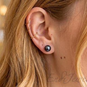May include: A single dark gray pearl stud earring worn in the earlobe, accompanied by three thin gold-toned hoop earrings positioned along the upper cartilage. The pearl has a smooth, lustrous finish, and the hoops include two plain bands and one twisted design.