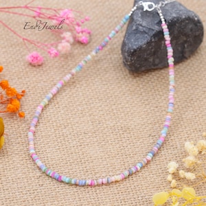 May include: A delicate necklace featuring small, colorful beads in pastel shades of pink, blue, yellow, and green. The beads are strung together with a silver clasp. The necklace is displayed on a neutral-toned surface with floral accents.