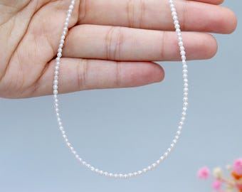 May include: A delicate white pearl necklace. The necklace is made up of many small, round pearls strung together. The pearls are a bright white colour, creating a simple, elegant design. The necklace is displayed against a light blue background.