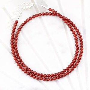 May include: A delicate necklace featuring small, round, reddish-brown beads. The beads are strung together, forming a double strand. The necklace has a silver-colored clasp and extender chain. The jewelry is displayed on a white surface.