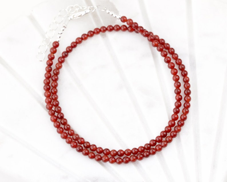 May include: A delicate necklace featuring small, round, reddish-brown beads. The beads are strung together, forming a double strand. The necklace has a silver-colored clasp and extender chain. The jewelry is displayed on a white surface.