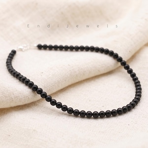 Puede incluir: Un delicado collar de cuentas negras con un cierre plateado, exhibido sobre una tela color crema. El collar est&aacute; compuesto por peque&ntilde;as cuentas negras redondas. El nombre de la marca "Endi Jewels" est&aacute; impreso en letras grises finas arriba.