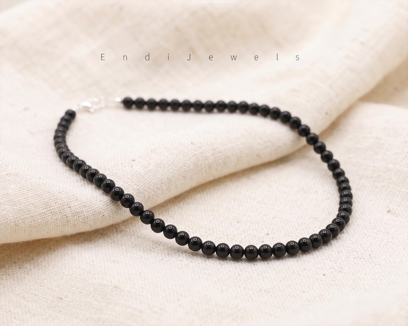 Puede incluir: Un delicado collar de cuentas negras con un cierre plateado, exhibido sobre una tela color crema. El collar est&aacute; compuesto por peque&ntilde;as cuentas negras redondas. El nombre de la marca "Endi Jewels" est&aacute; impreso en letras grises finas arriba.