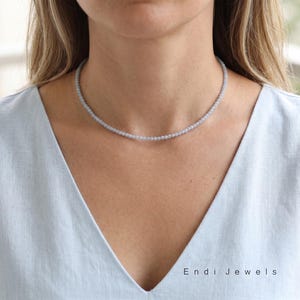 Pode incluir: Um delicado colar de contas azul claro. O colar é composto por pequenas contas redondas e repousa suavemente sobre o decote. A imagem também exibe o texto "Endi Jewels".